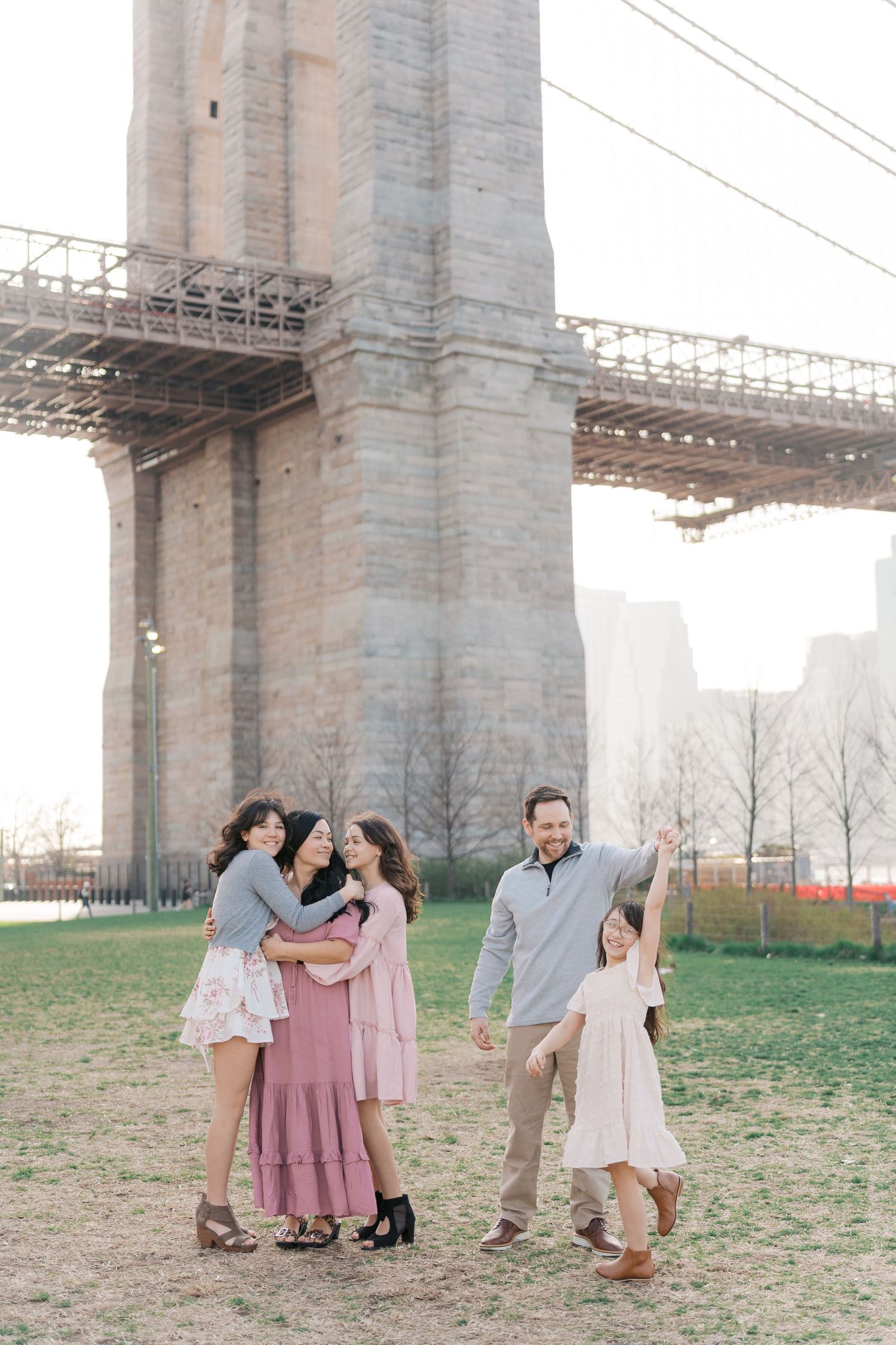 Family of five posing under the Brooklyn Bridge in a park, wearing pastel outfits on a bright day.