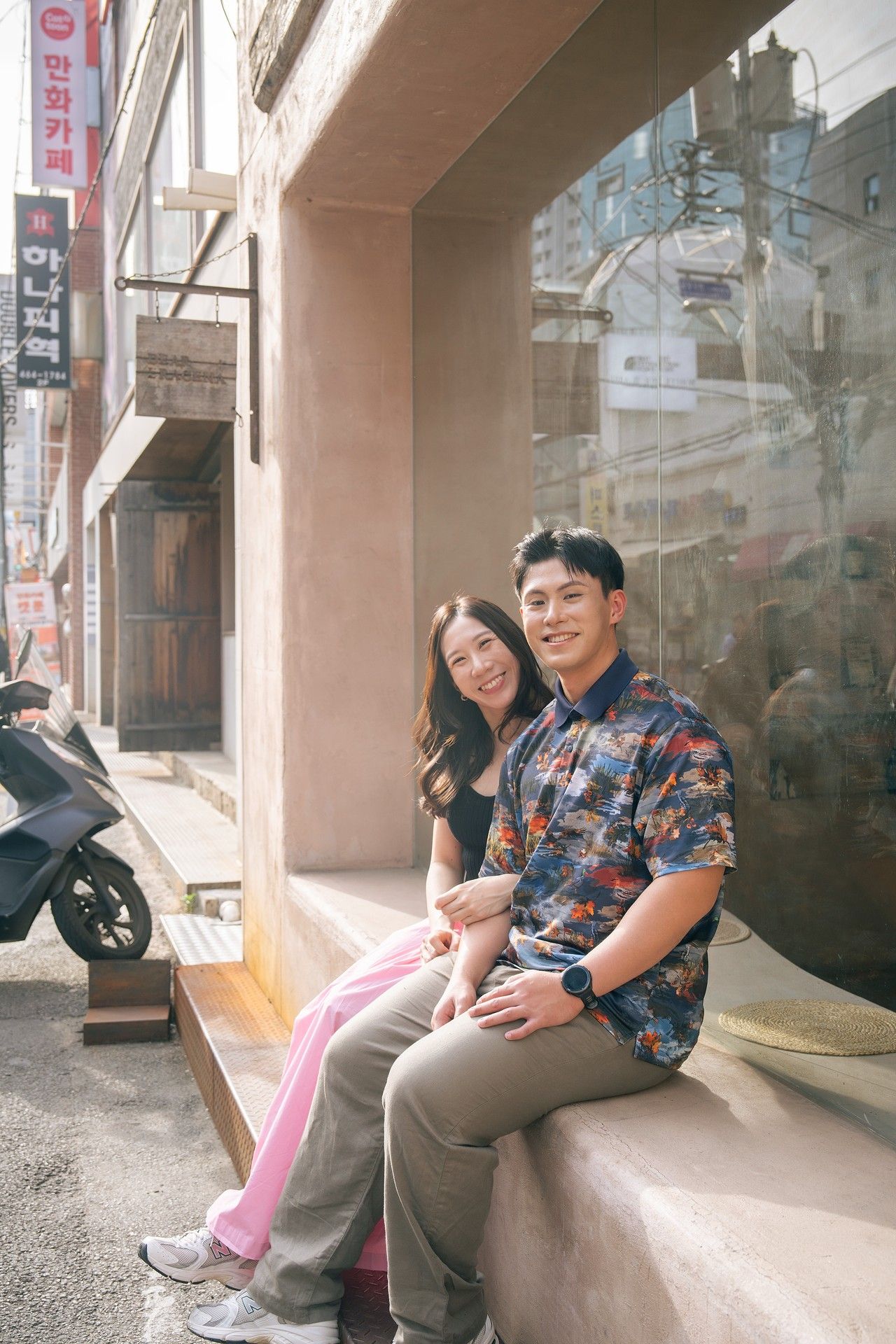 A smiling couple sits together on a bench outside a shop on a Korean street, wearing casual colorful outfits.