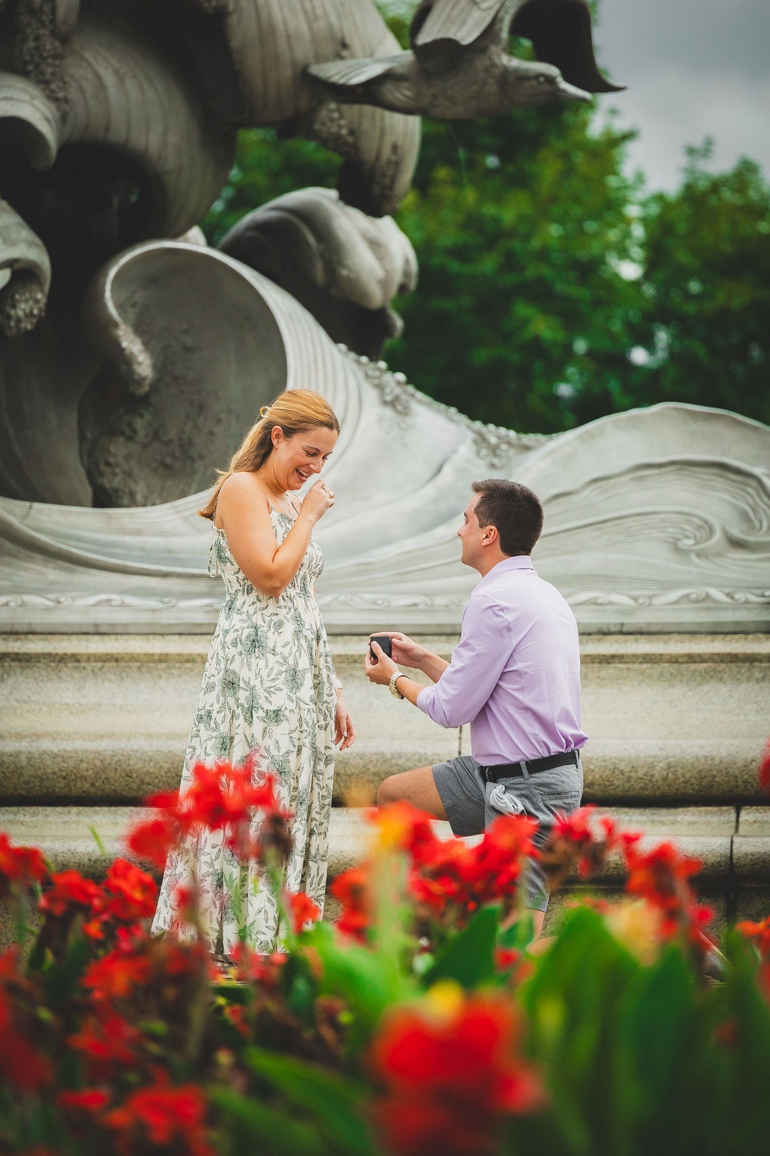 Man proposes to woman on one knee near ornate fountain with red flowers in foreground at the navy merchant marine memorial