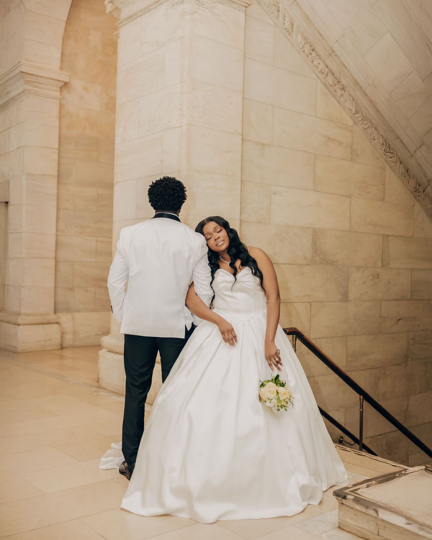 Bride in white ball gown holds bouquet beside groom in white tuxedo at stone venue.