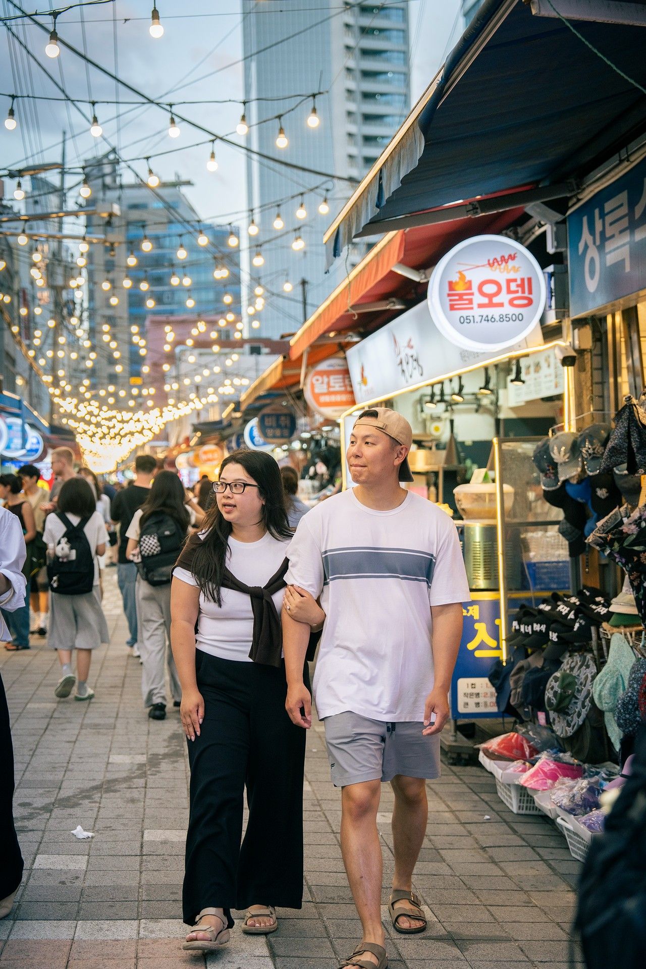 A couple walks hand in hand through a bustling Korean street market with string lights and shop signs at dusk.