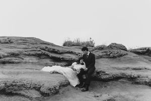 Black and white photo of a bride and groom sitting together on rocky terrain outdoors.