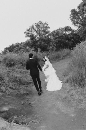 Black and white photo of a bride and groom walking hand in hand along a scenic outdoor path surrounded by lush greenery.