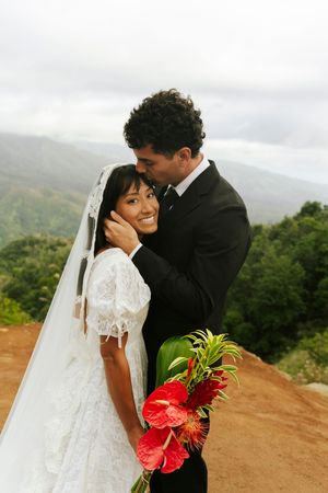 Bride in white lace gown holding red tropical bouquet embraced by groom in black suit on scenic mountain overlook.