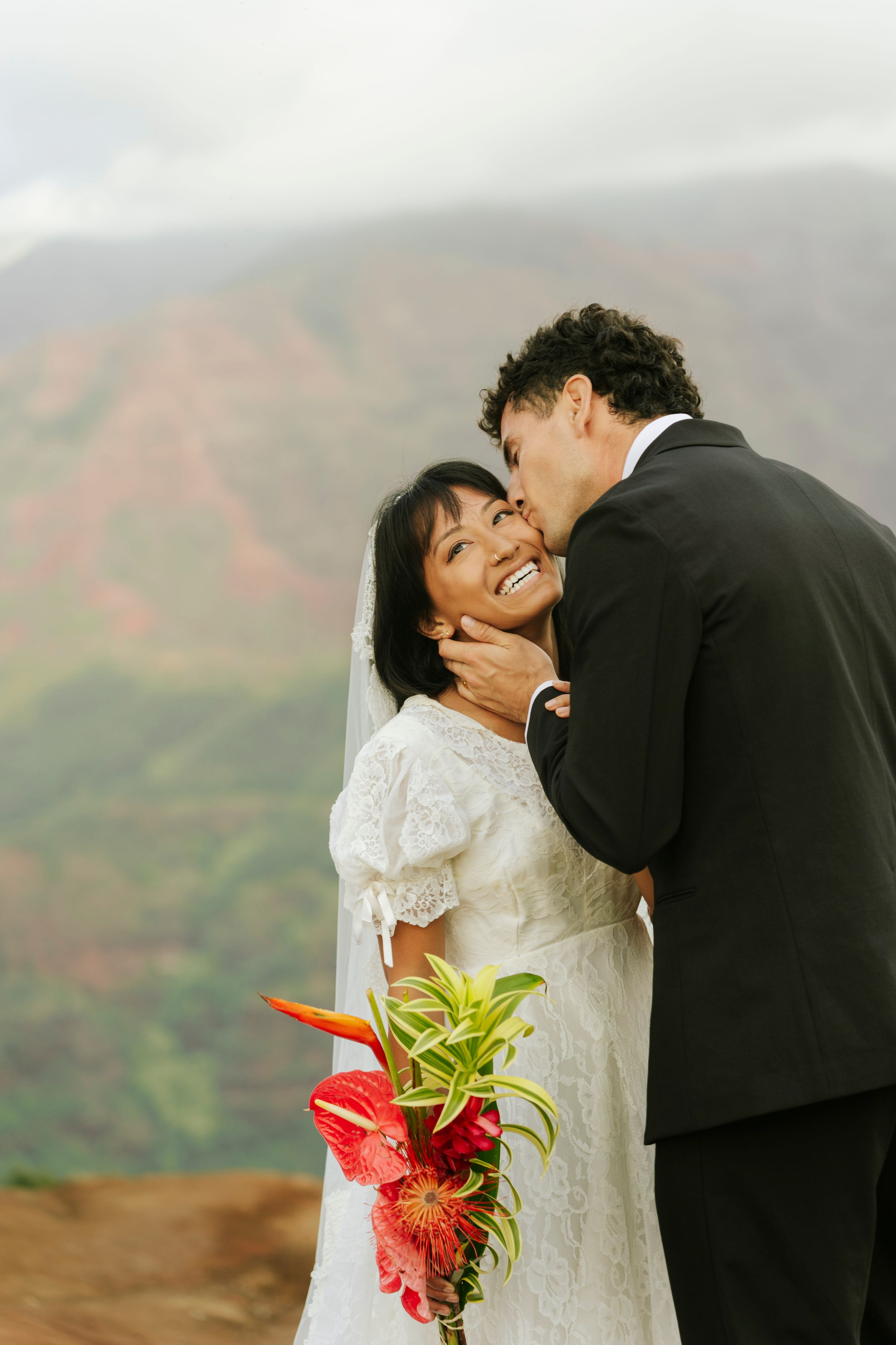 Bride smiling as groom kisses her cheek on a scenic mountain overlook, holding tropical red bouquet.