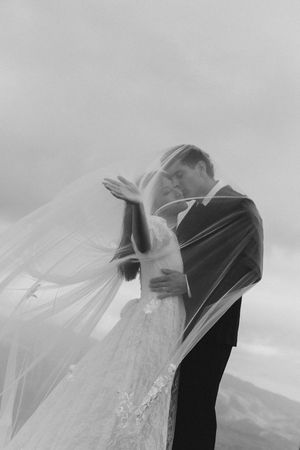 Black and white wedding photo of bride and groom embracing under flowing veil outdoors.