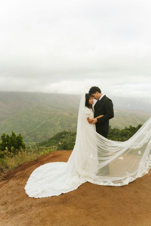Wedding couple embracing with flowing lace veil on mountain overlook with lush green valley backdrop.