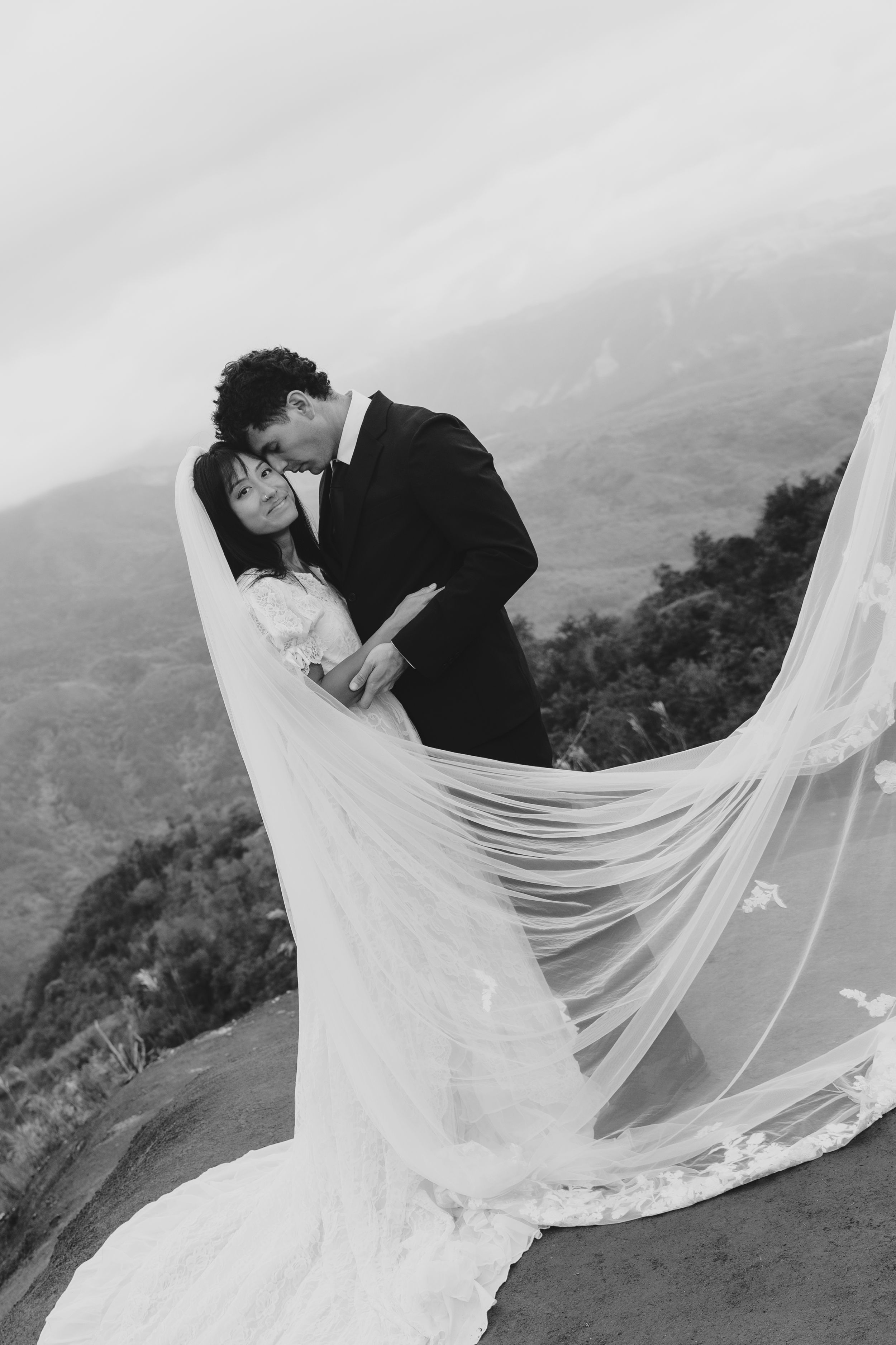 Black and white photo of bride and groom kissing on mountain overlook with veil flowing.