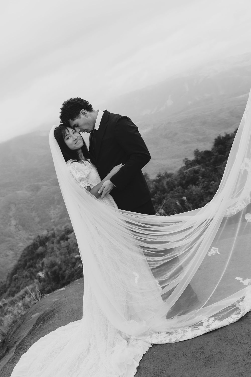 Black and white photo of bride and groom kissing on mountain overlook with veil flowing.