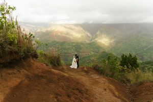 Couple in wedding attire kissing on a scenic mountain overlook with lush green valleys and misty peaks in Kauai, Hawaii.