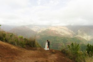 Couple in wedding attire embracing on a scenic mountain overlook with lush green valleys and misty peaks in the background.