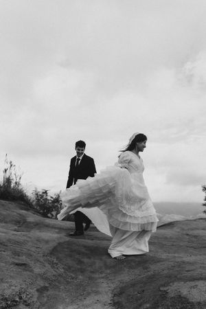 Black and white photo of a bride in flowing gown and groom in suit walking on a hillside on their wedding day.
