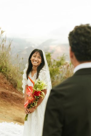 Radiant bride in lace dress holding tropical bouquet smiles at groom during outdoor mountain elopement.