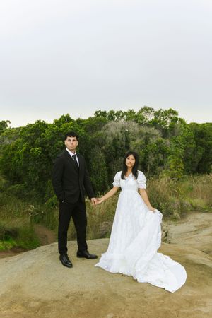Bride in white gown holds groom's hand in black suit on rocky outcrop with lush green trees behind them.