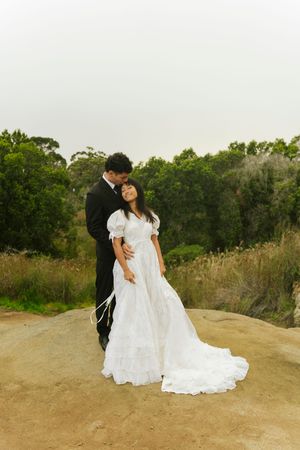 Groom in black suit embraces bride in white wedding dress on rocky terrain surrounded by green bushland.