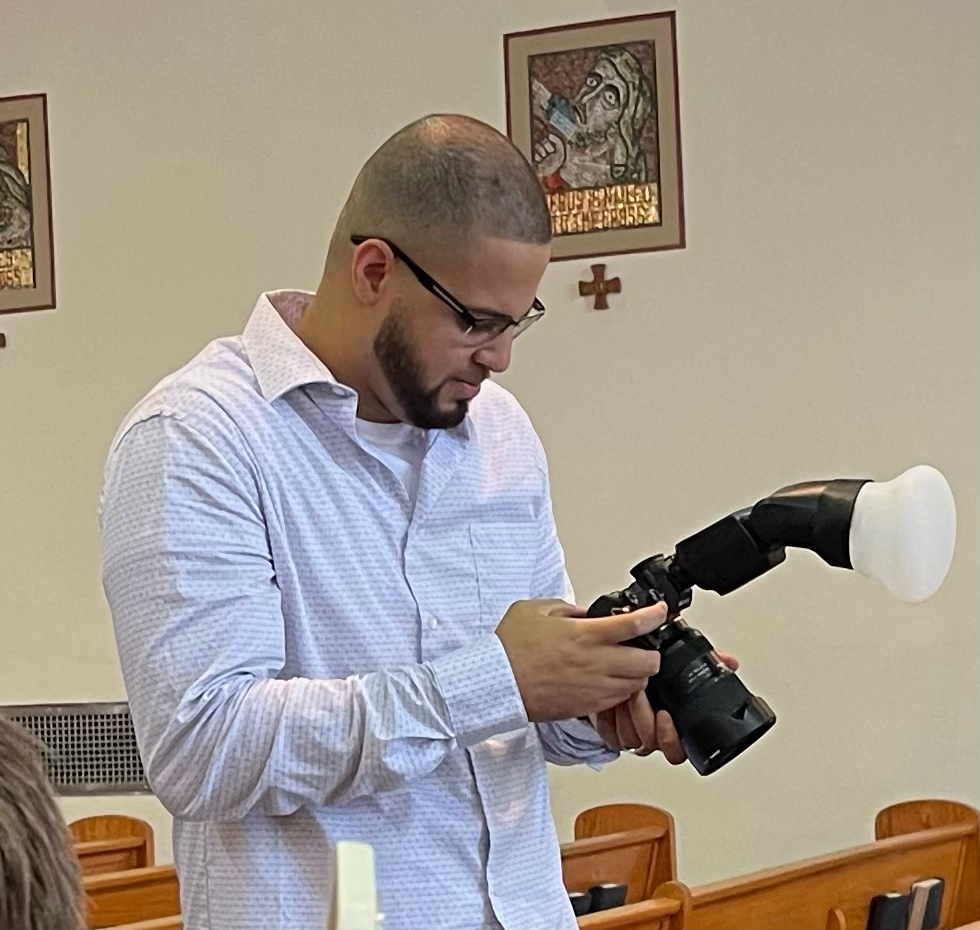 Photographer with camera and flash diffuser stands between church pews during indoor religious ceremony.