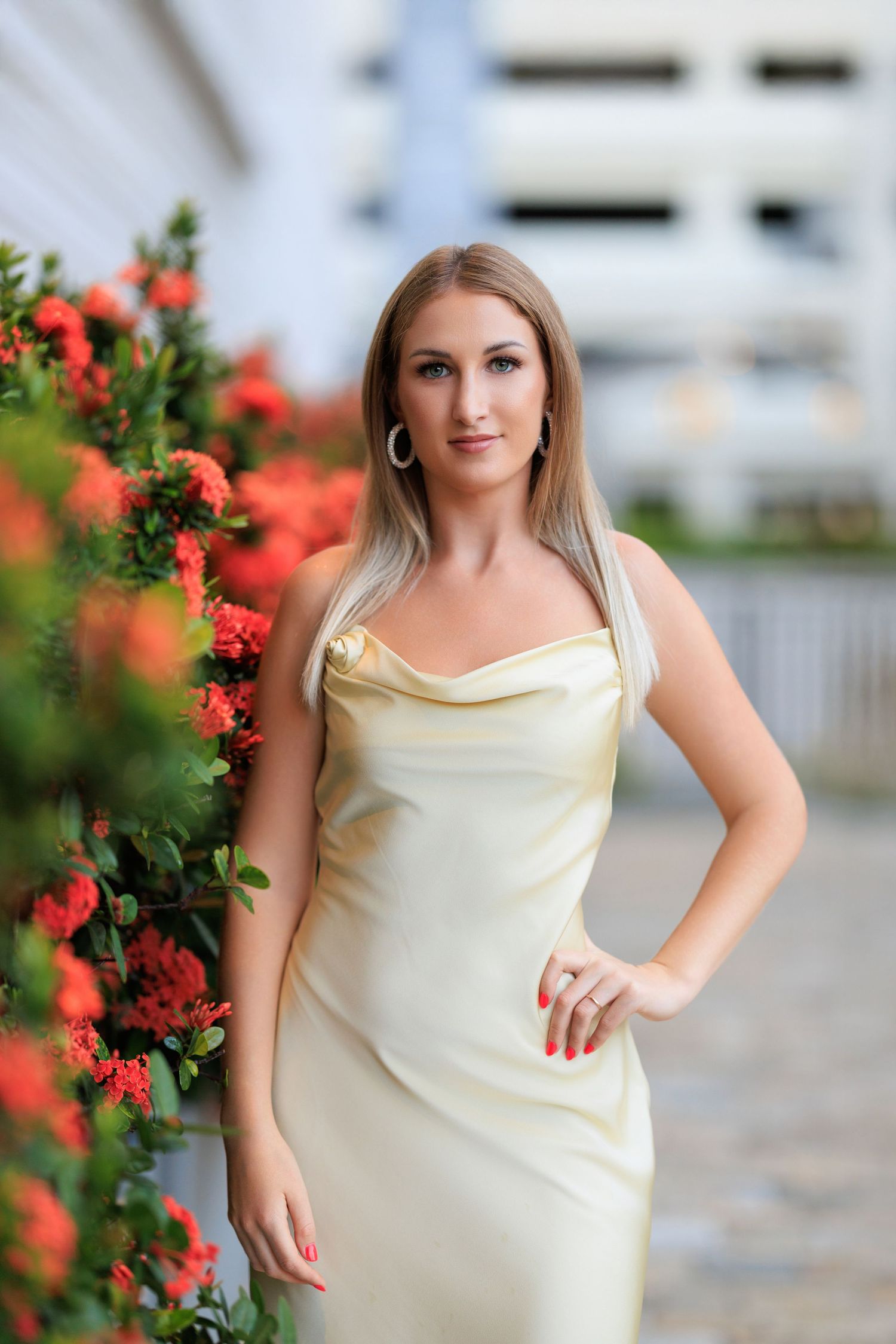 Elegant blonde woman in a gold satin dress posing beside vibrant red flowers outdoors.