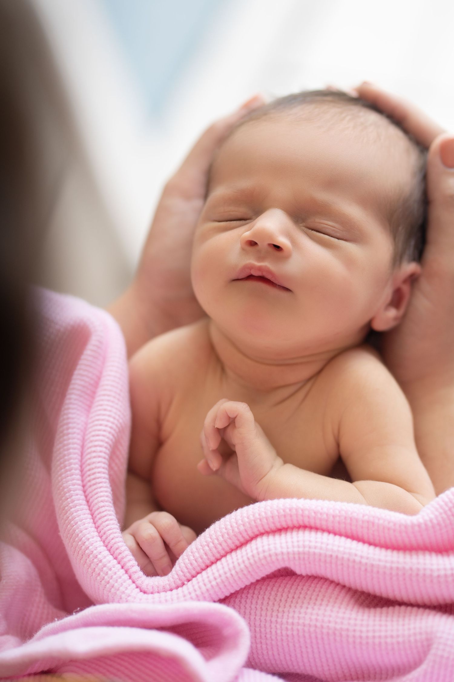 Newborn baby sleeping peacefully wrapped in a pink blanket while being cradled in caring hands.