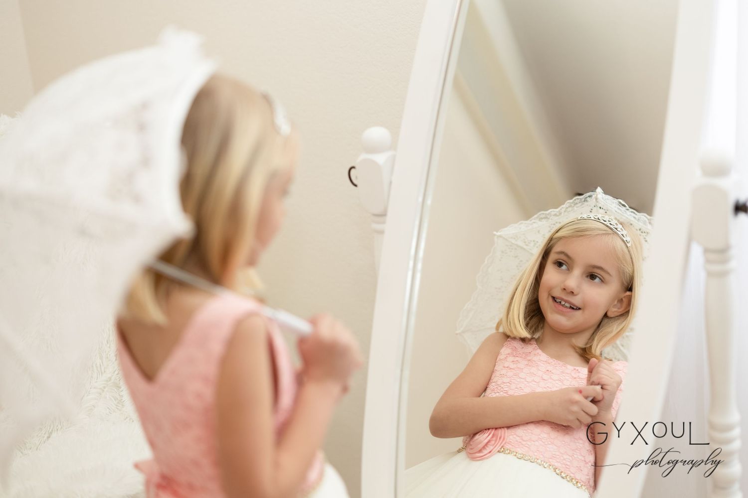 Young girl in pink dress and tiara smiles at her reflection in a tall white mirror, holding a white umbrella.