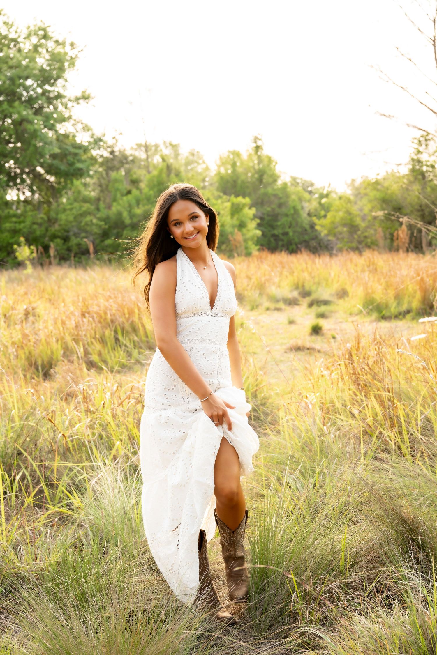 A smiling woman in a pink dress stands in golden tall grass surrounded by pine trees in a sunny outdoor setting.