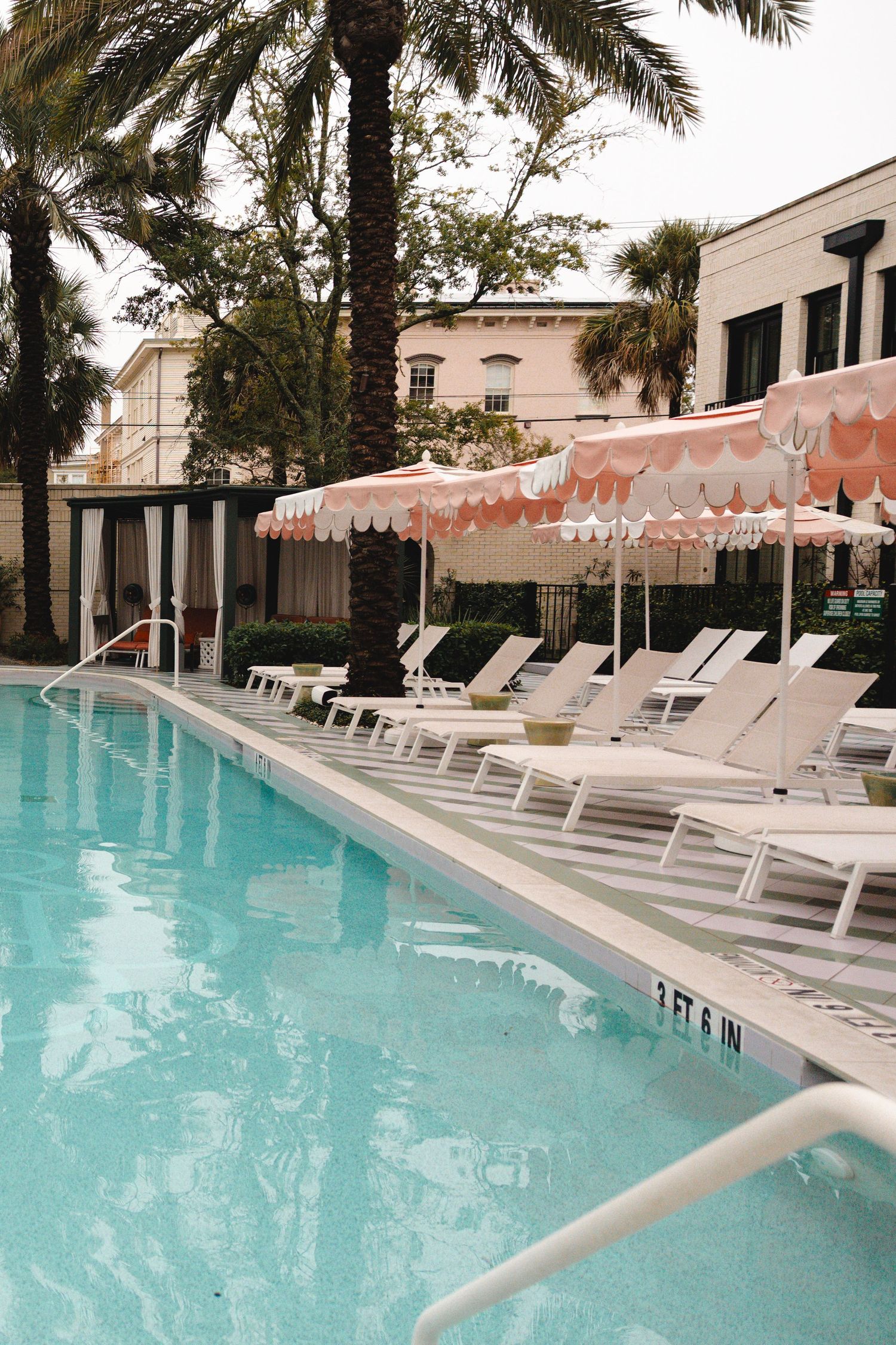 Luxury hotel pool with white lounge chairs, pink scalloped umbrellas, and palm trees on a cloudy day.