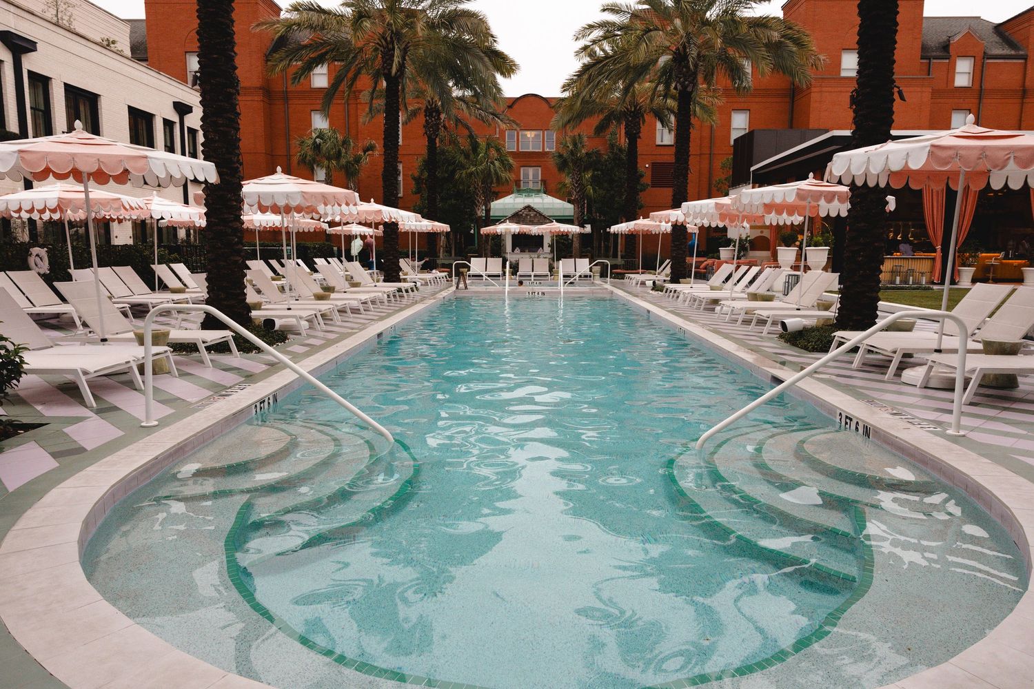 Resort swimming pool with curved steps, striped umbrellas, lounge chairs, and palm trees beside a terracotta-colored hotel building.