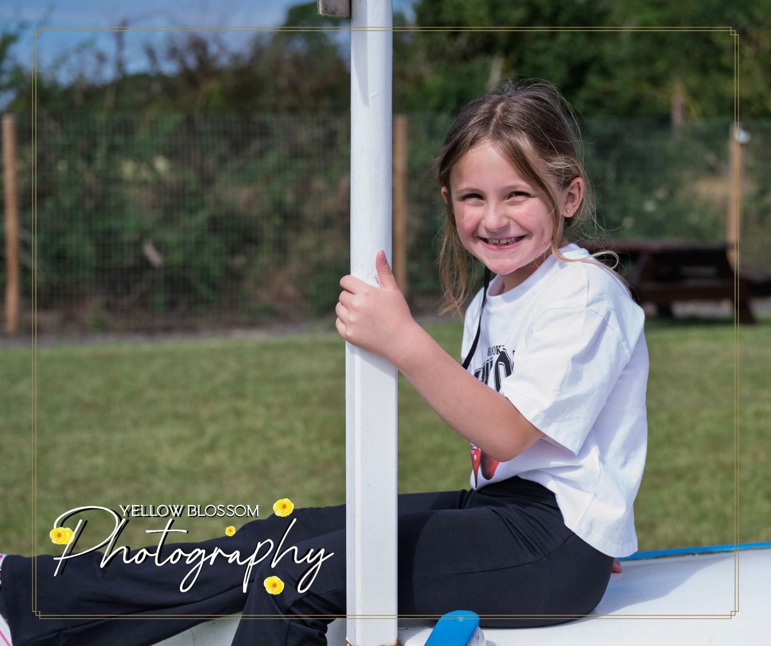 Young karate student in white uniform sits on ground next to white fence in outdoor training area.