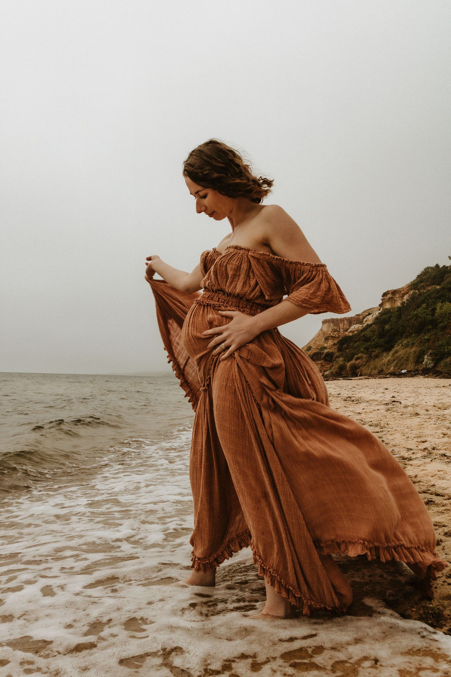 Woman in flowing brown dress standing in ocean waves near rocky coastline.