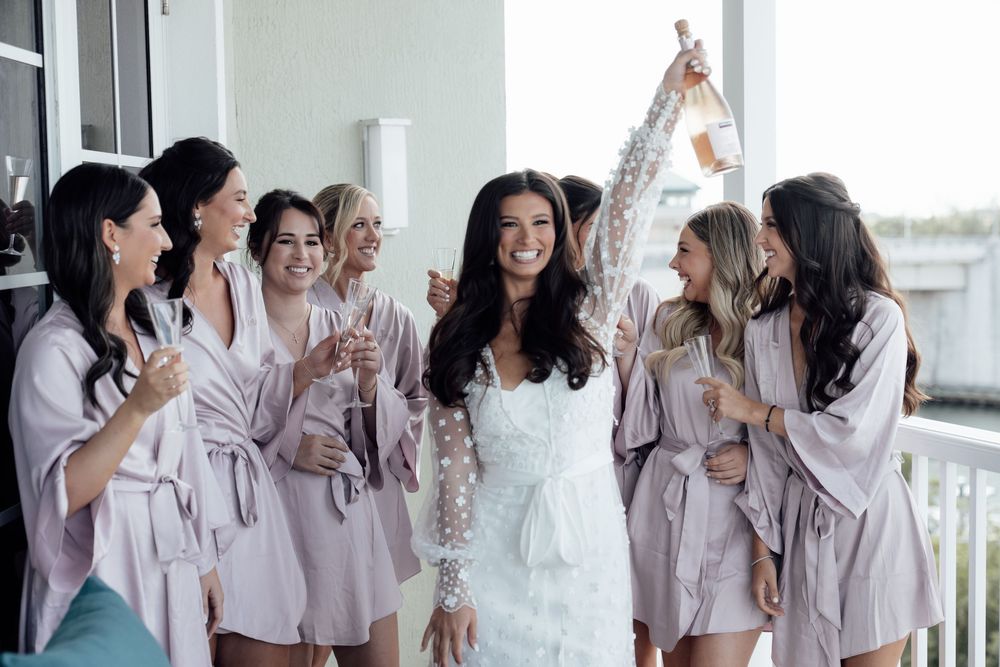 Bridesmaids in matching pink silk robes laughing and celebrating together in a bright room during wedding preparations.
