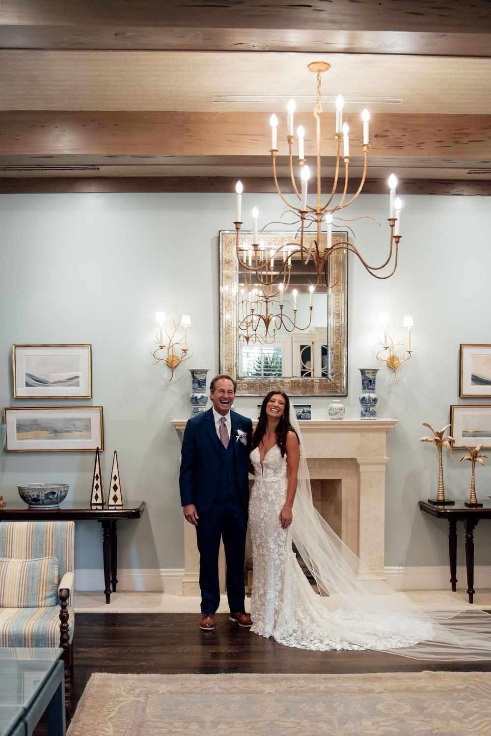 A couple stands in an elegant room with chandeliers, wooden beams, and a stone fireplace creating a luxurious wedding setting.