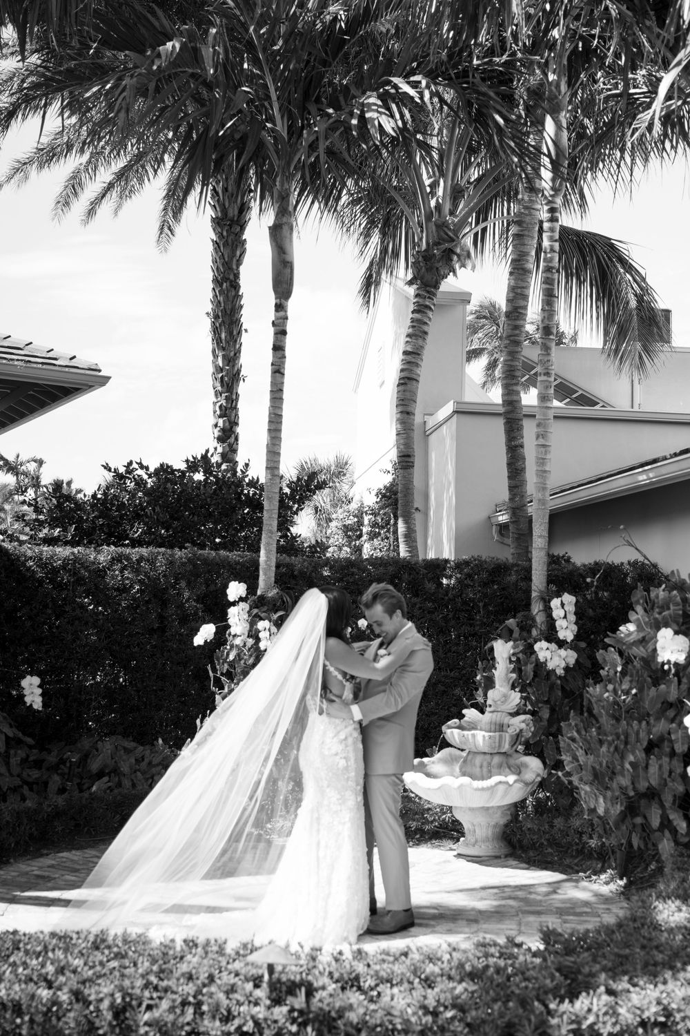 Wedding couple embraces in a tropical garden with palm trees and elegant white architecture in the background.