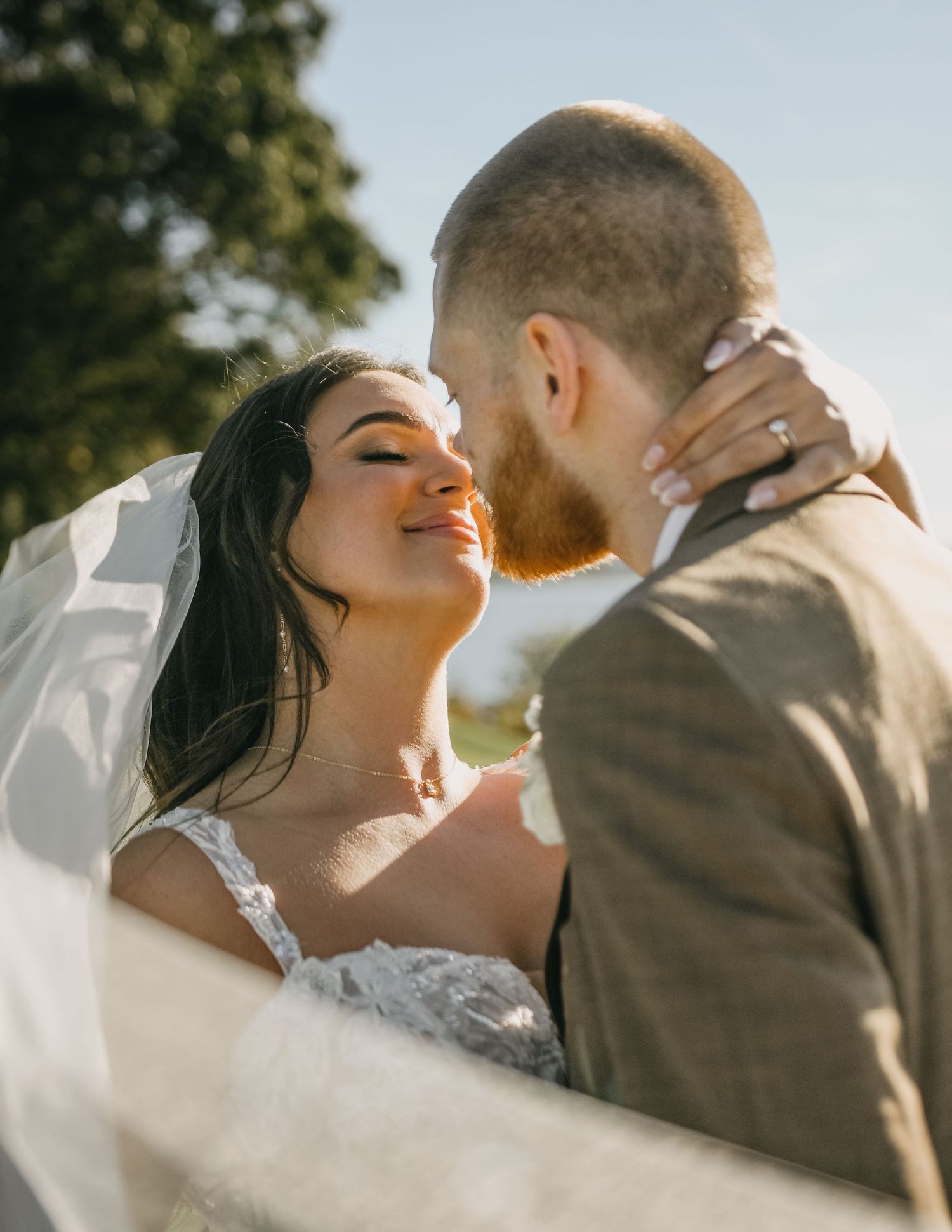 Romantic bride and groom sharing a kiss outdoors, bride in lace gown with veil flowing in sunlight.