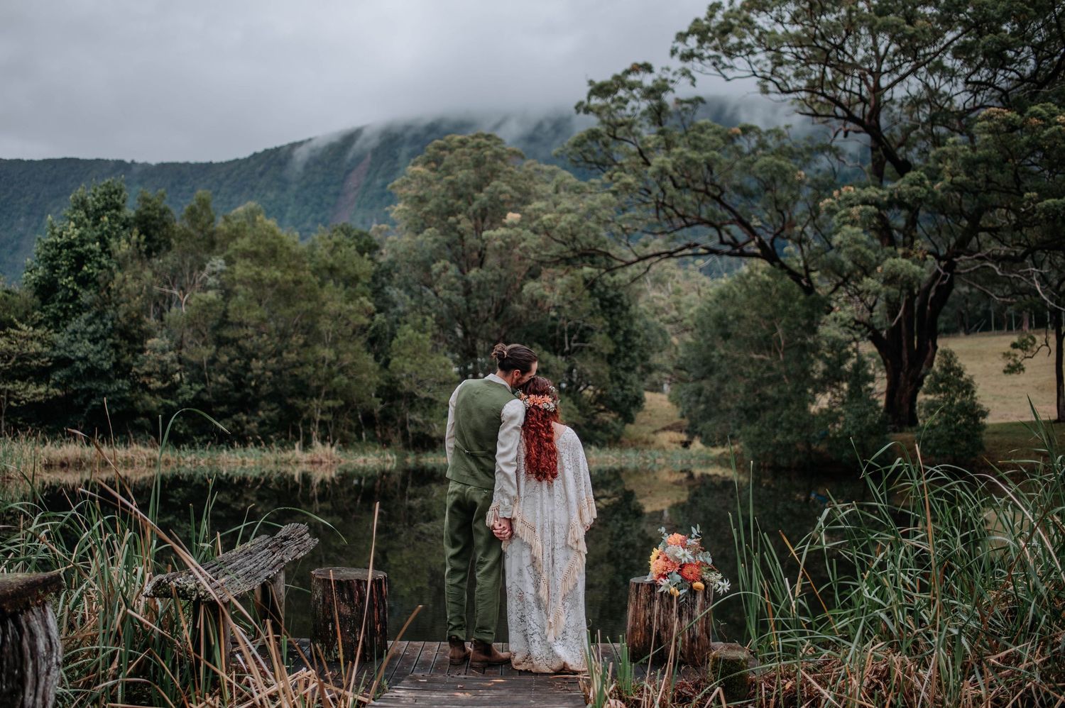 Couple stands on wooden dock by misty mountain lake surrounded by lush trees and tall grasses in moody lighting.