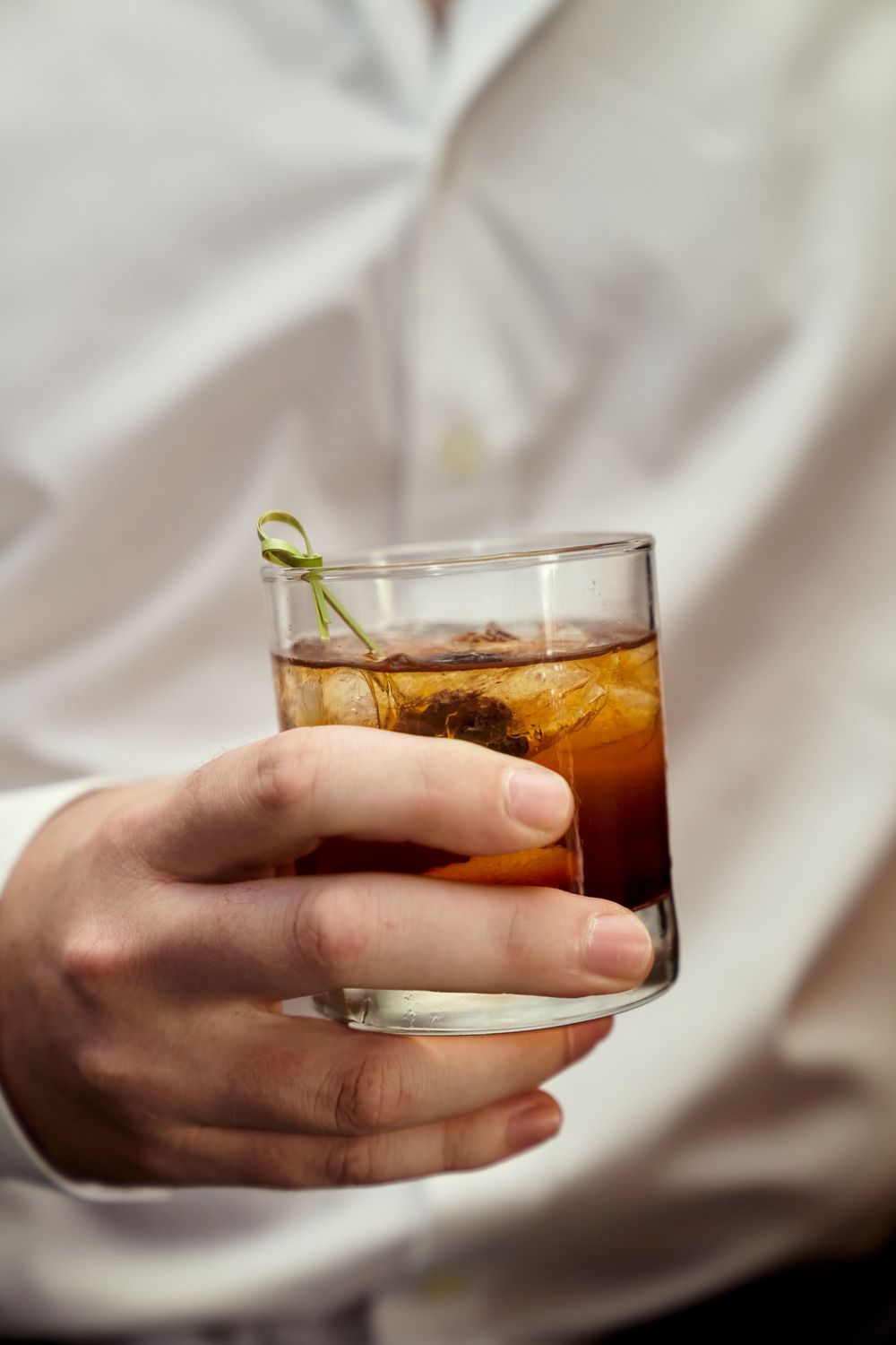 A glass of whiskey or bourbon cocktail with ice being held against a white fabric background. Engagement photoshoot