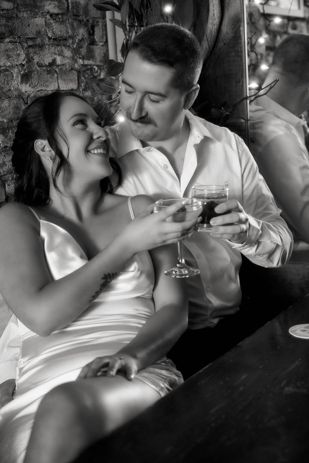 A romantic black and white photo of a couple toasting with wine glasses in an intimate bar setting. Engagement photoshoot