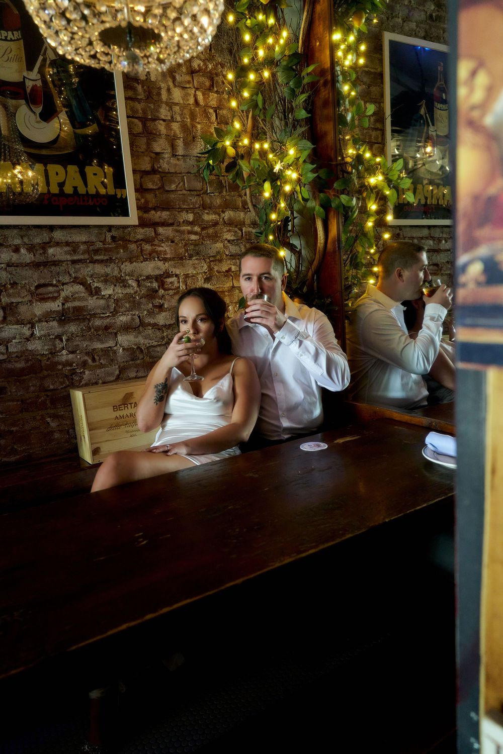 A couple sits at a rustic wooden table in a restaurant with exposed brick walls and twinkling fairy lights overhead. Engagement photoshoot