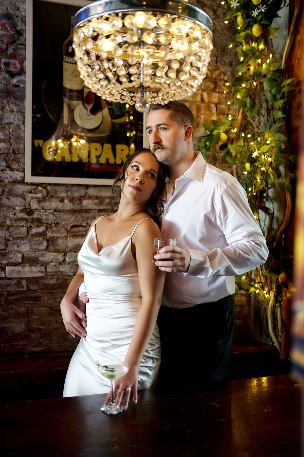 A couple dances intimately beneath an ornate crystal chandelier in an atmospheric venue with brick walls and twinkling lights. Engagement