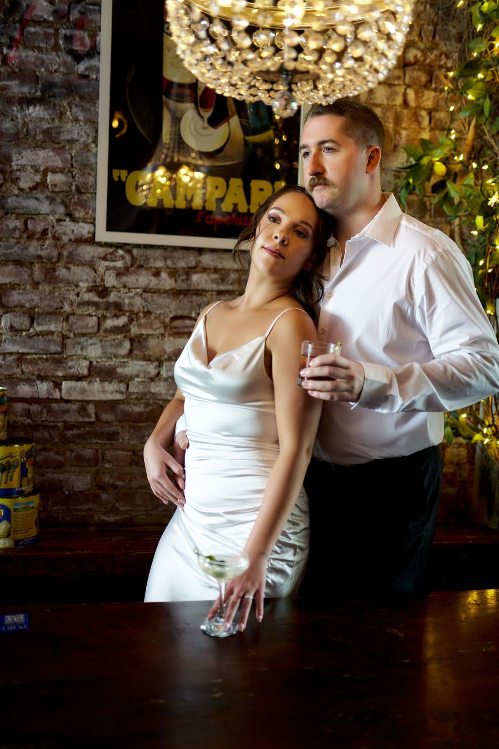 A couple shares an elegant moment beneath a crystal chandelier in a rustic brick-walled venue during a celebration. Engagement photoshoot