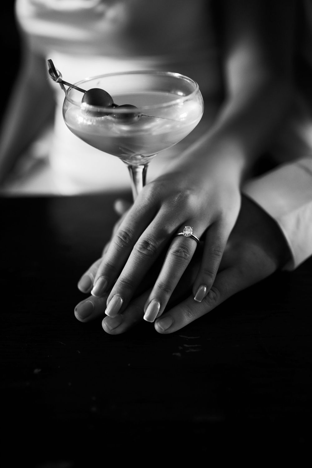 Elegant black and white close-up of a martini glass with olive garnish and a hand wearing rings resting on dark surface. Engagement photosho
