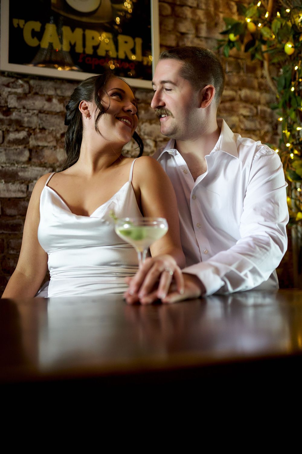 Romantic couple sharing intimate moments at a dimly lit bar with holiday lights and brick walls in background. Engagement photoshoot