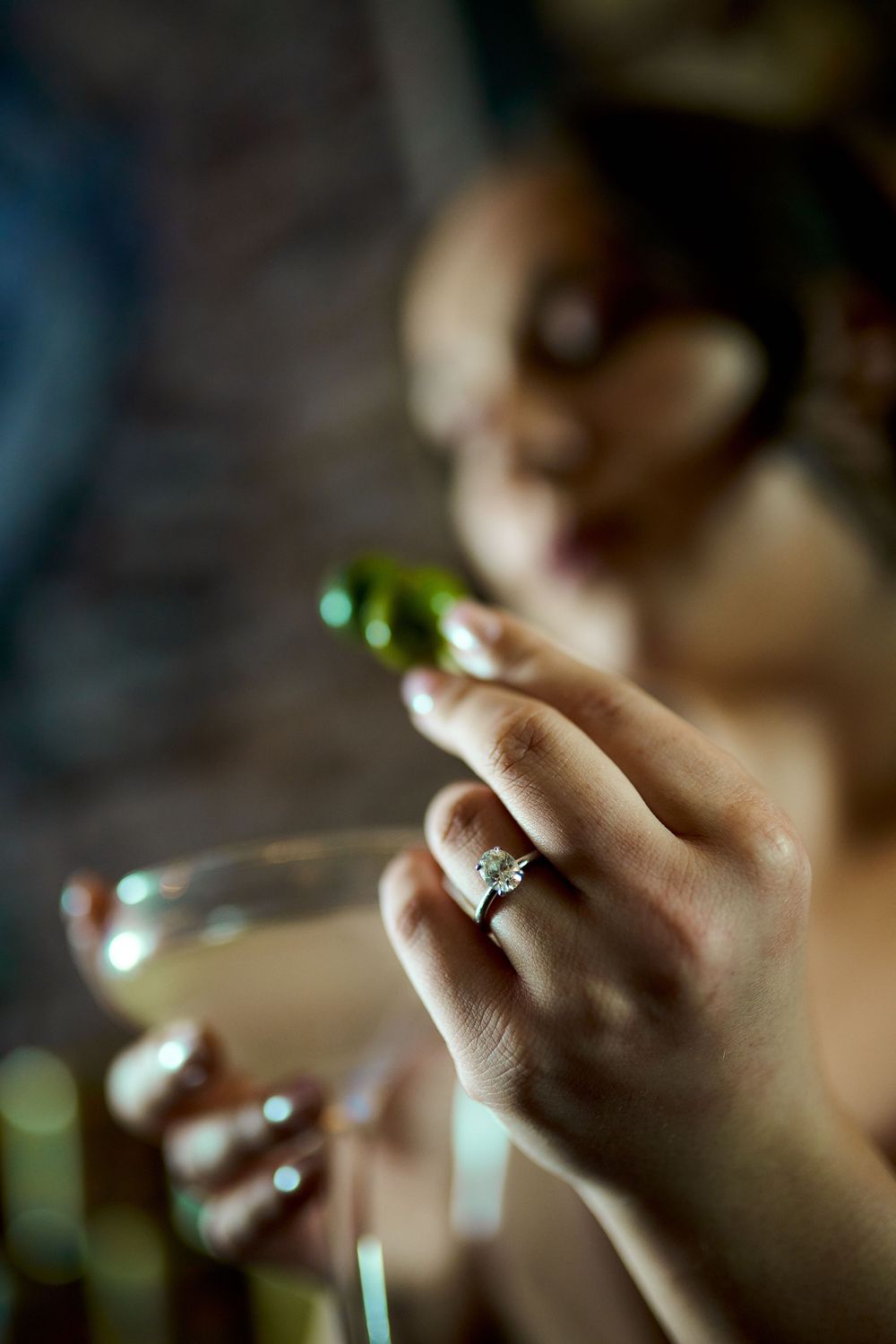 Close-up shot of a hand wearing an engagement ring while holding a small bottle in dim lighting. Engagement photoshoot