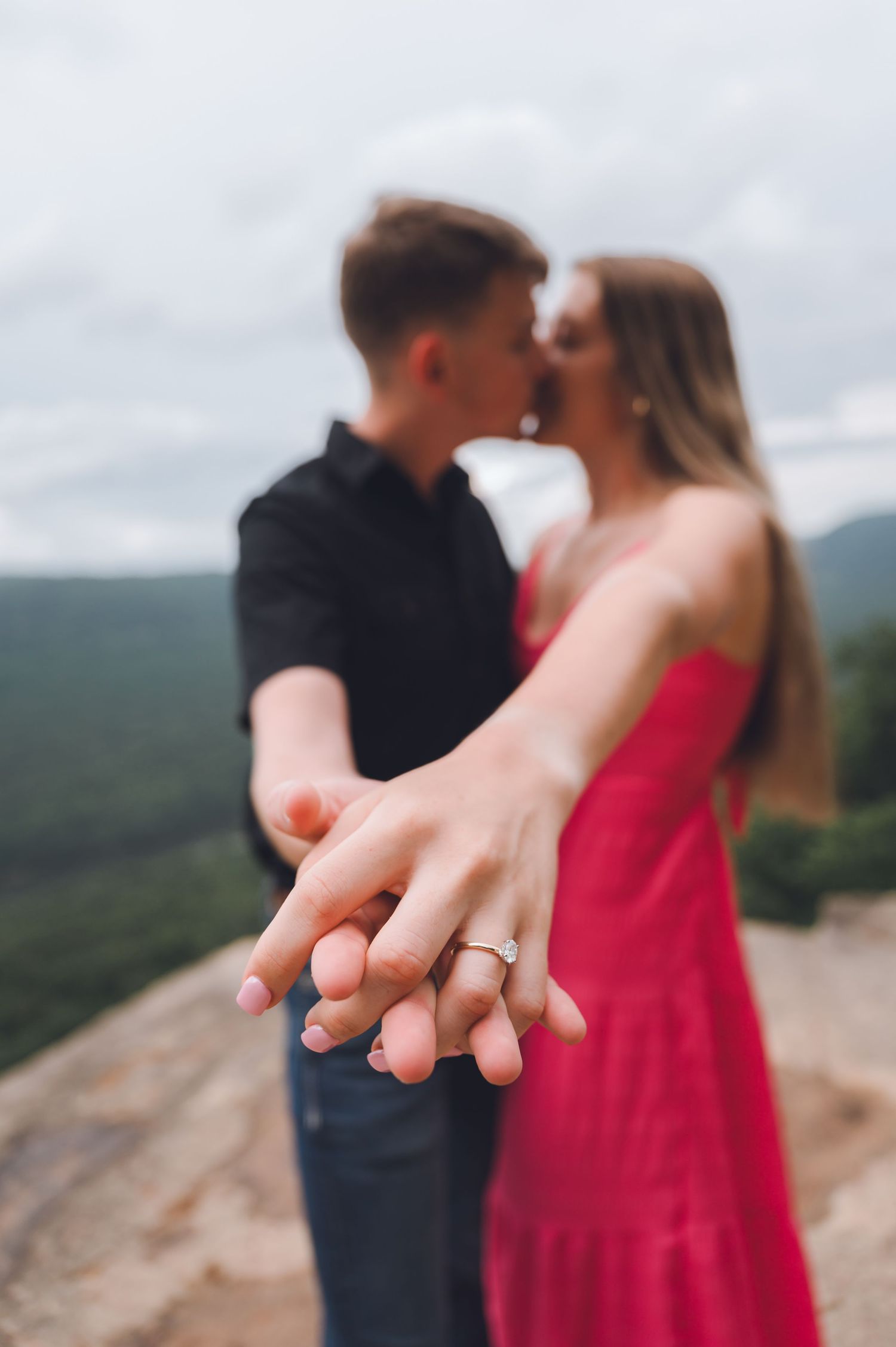A couple in a red dress and black shirt hold hands while sharing an intimate moment on a mountain overlook.