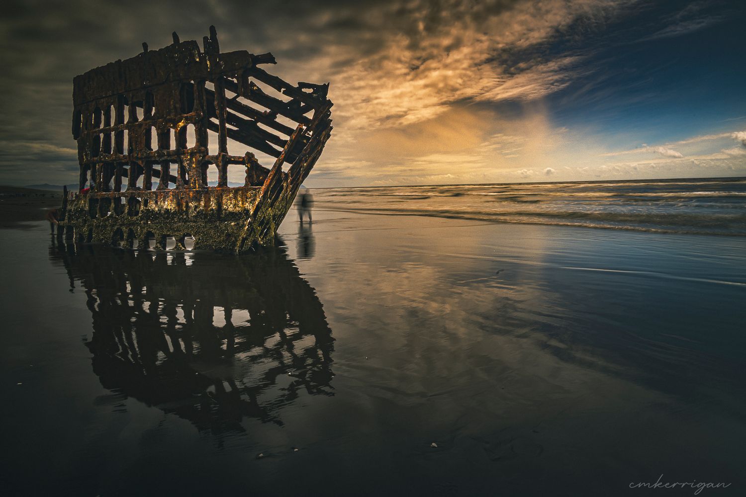 Shipwreck remains on a beach at sunset with dramatic golden clouds reflected in wet sand.