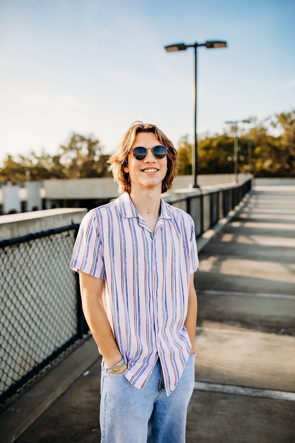 A person wearing a striped button-down shirt and jeans poses outdoors on a bright sunny day with railing in background.