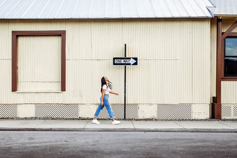Rear view of a person in a blue dress and white sneakers walking next to the exterior wall of a beige building.