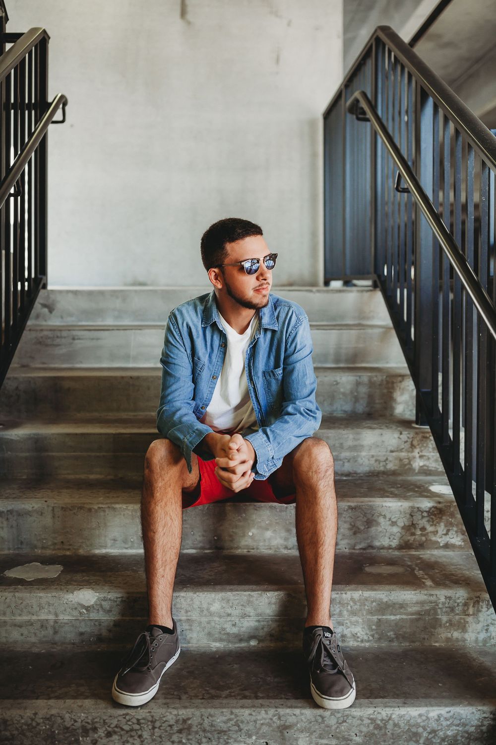 Person in denim shirt and sunglasses sits on concrete steps in urban setting looking contemplative.