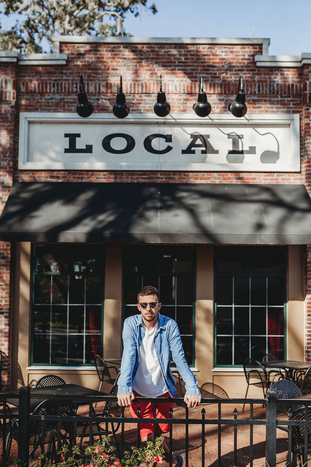 A stylish man in sunglasses and blue blazer leans on a railing outside a local cafe with brick storefront.