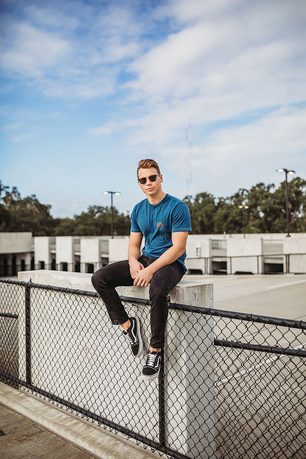Person in blue shirt poses casually on industrial rooftop railing against cloudy sky.