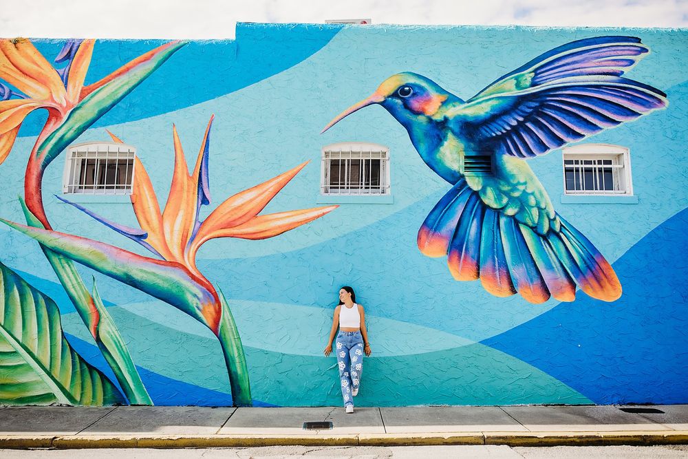 A vibrant street art mural features colorful hummingbirds and tropical flowers against a bright blue background.