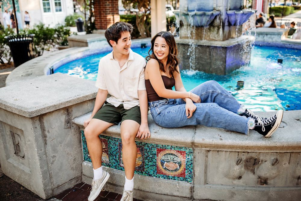 Two people sit casually together on a stone fountain ledge in front of blue pool water and ornate architecture.